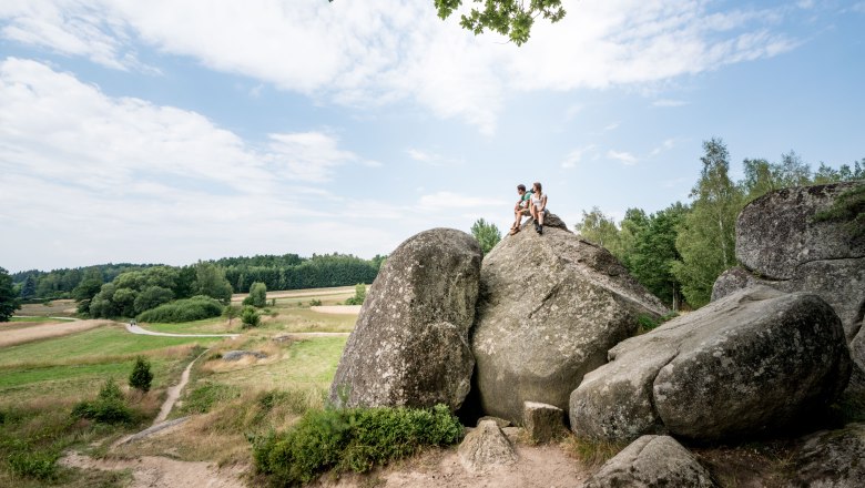 Zwei Personen sitzen auf einem großen Felsen in einer ländlichen Landschaft mit Wiesen und Bäumen.