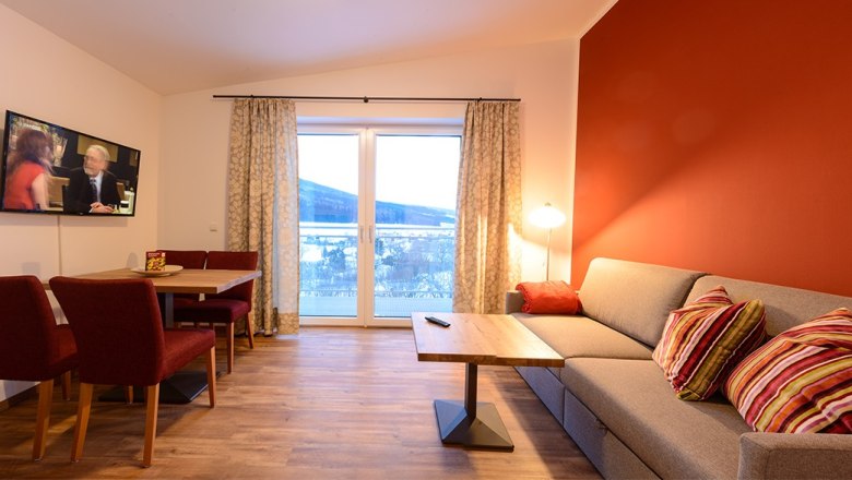 Gemütliches Wohnzimmer mit roter Akzentwand, grauem Sofa, Holztisch, Fernseher und großem Fenster mit Vorhängen.