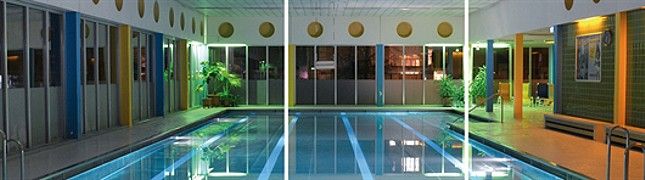 Innenansicht eines beleuchteten Hallenbads mit Schwimmbecken und Pflanzen.