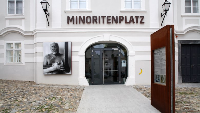 Eingang zum Forum Frohner am Minoritenplatz mit Kunstwerk und Infotafel.