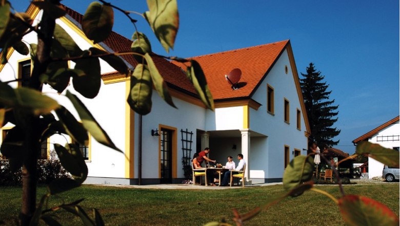 Ein wei&szlig;es Haus mit rotem Dach, umgeben von gr&uuml;nem Rasen und B&auml;umen. Menschen sitzen an einem Tisch im Freien.