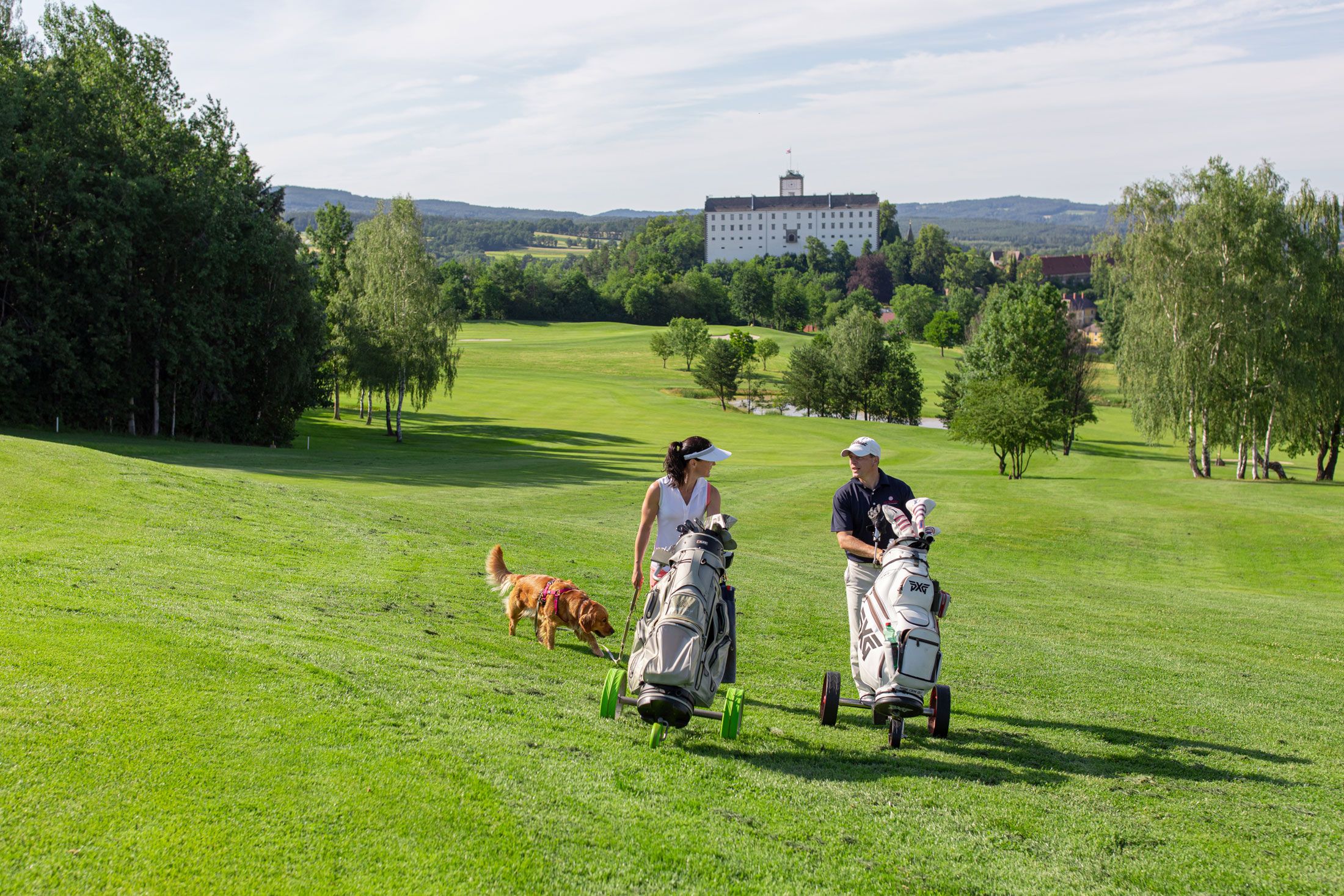 Zwei Golfer mit Hund auf grünem Golfplatz, Schloss im Hintergrund.