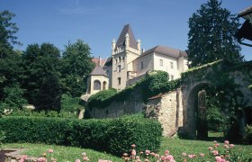 Ein Schloss mit T&uuml;rmen und einer Steinmauer, umgeben von gr&uuml;nen B&auml;umen und bl&uuml;henden Blumen im Vordergrund.