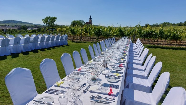 Lange, festlich gedeckte Tafel im Freien mit wei&szlig;en St&uuml;hlen und Tischdecken, umgeben von Weinreben und einer Kirche im Hintergrund.