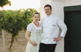 Ein Paar in traditioneller Kleidung steht vor einem wei&szlig;en Geb&auml;ude neben einem Weinberg.