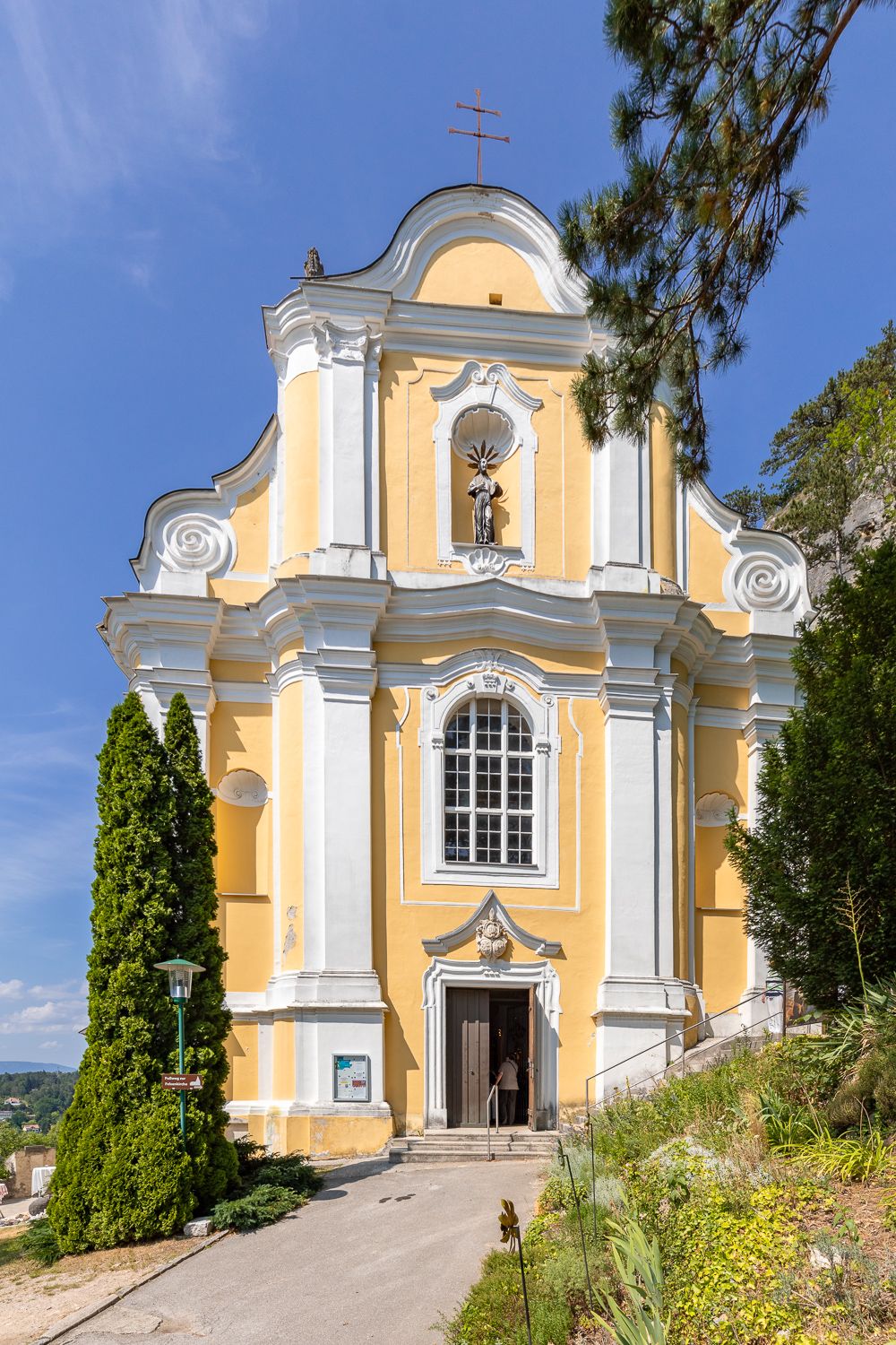 Gelbe Barockkirche mit weißen Säulen und einer Statue über dem Eingang, umgeben von Bäumen und blauem Himmel.
