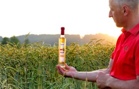 Ein Mann in rotem Hemd h&auml;lt eine Flasche Dinkelbrand in einem Getreidefeld bei Sonnenuntergang.