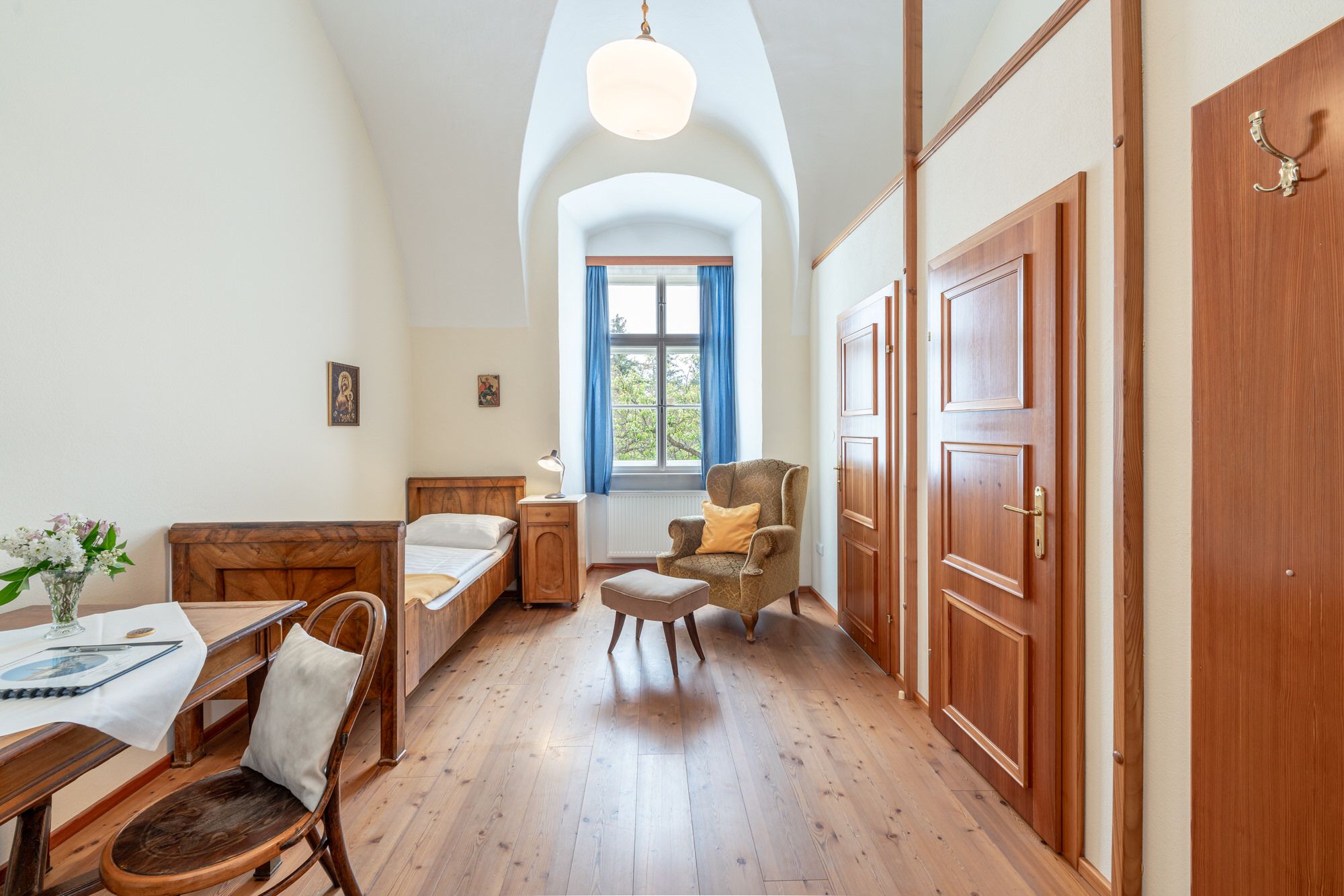 Einzelzimmer im Stift Altenburg mit Bett, Sessel, Tisch und Fenster mit blauen Vorhängen.