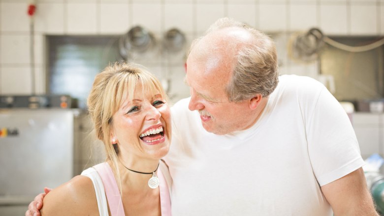 Zwei lachende Personen in einer K&uuml;che, eine Frau und ein Mann, in wei&szlig;er Kleidung.