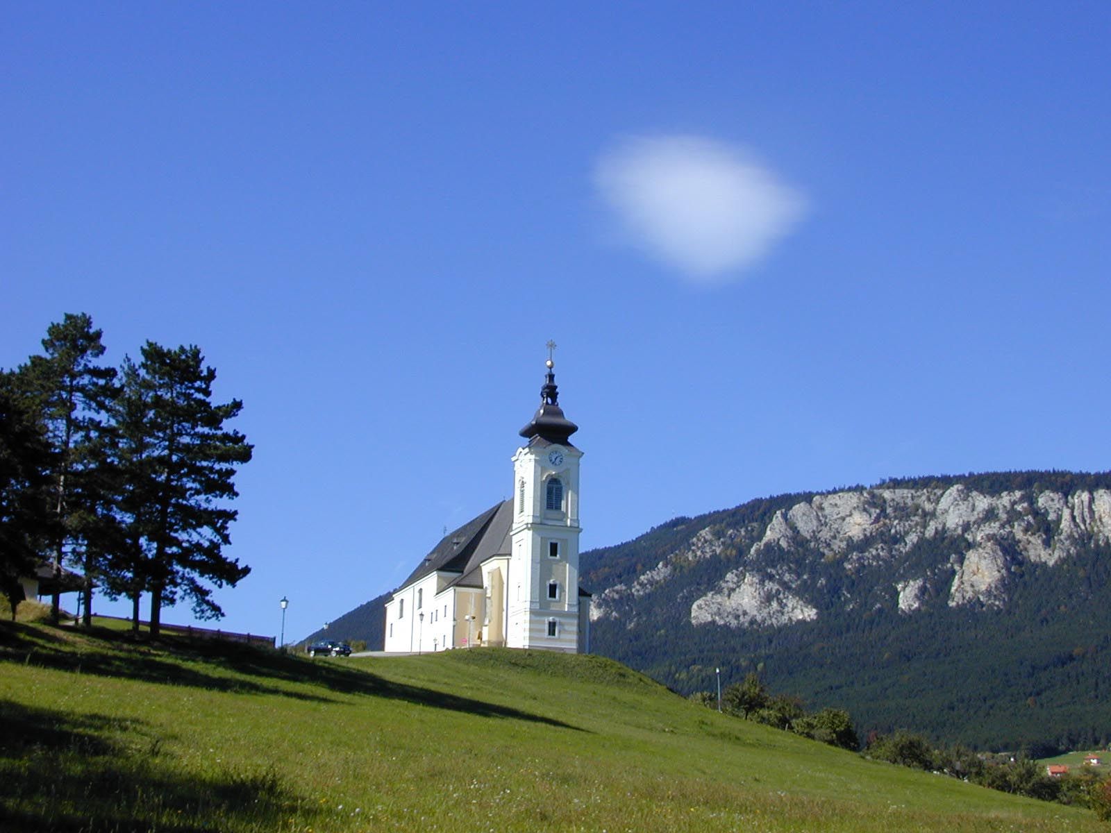 Kirche auf einem Hügel mit blauem Himmel und Bergen im Hintergrund.
