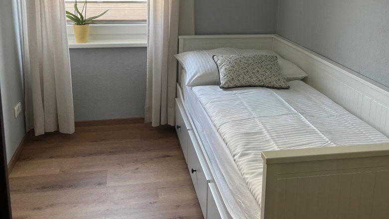 Kleines Schlafzimmer mit Einzelbett, wei&szlig;en Vorh&auml;ngen und einer Pflanze auf der Fensterbank.