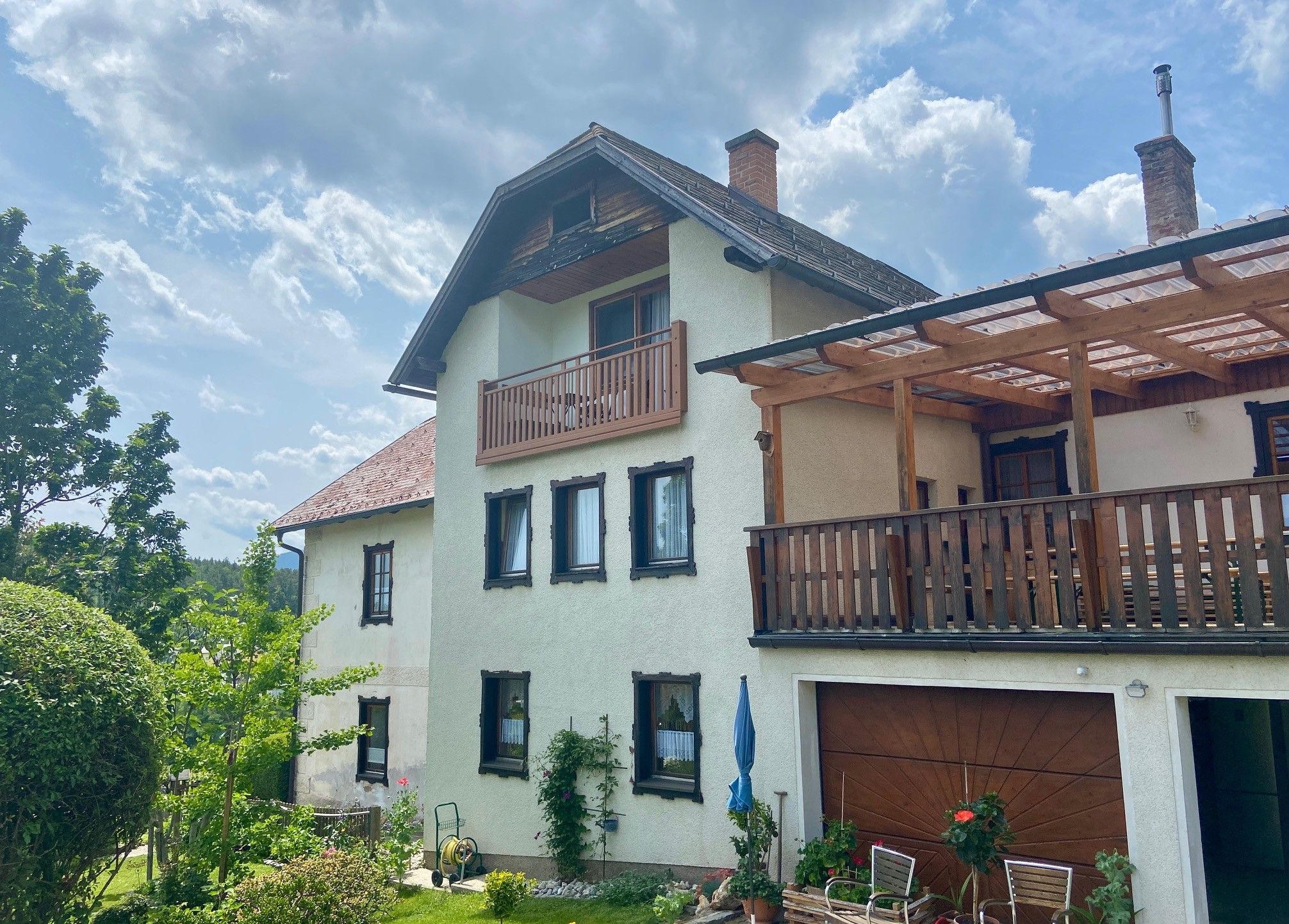 Ein zweistöckiges Haus mit Balkon und Terrasse, umgeben von Garten und Bäumen, unter einem bewölkten Himmel.