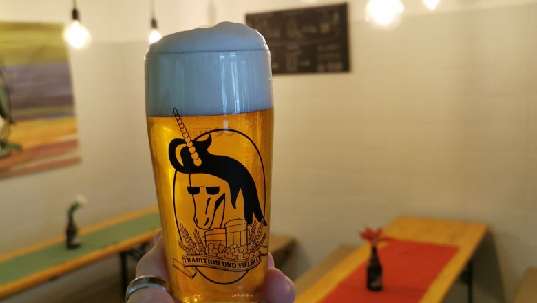 Ein Glas Bier mit Schaumkrone wird in einem Verkostungsraum mit Holztischen und Lichterketten gehalten.