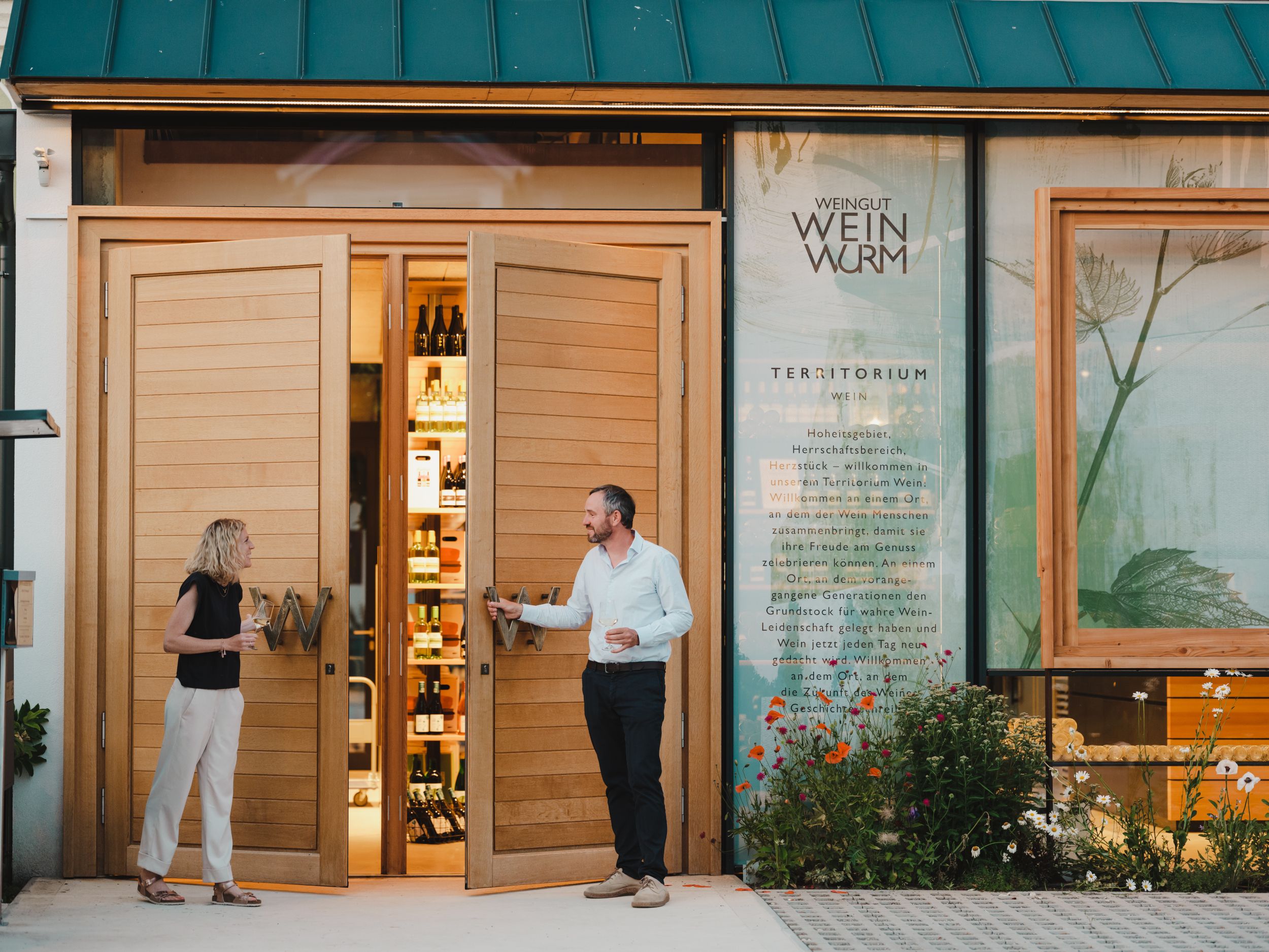 Zwei Personen stehen vor einem Weingut mit geöffneten Holztüren, die Flaschen im Inneren zeigen.