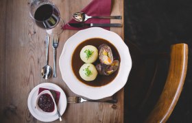 Ein Teller mit Rindsroulade und zwei Erdäpfelknödeln in Soße, daneben ein Glas Rotwein und Preiselbeeren auf einem Holztisch.