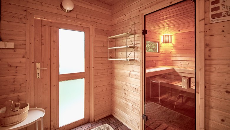 Innenansicht einer Sauna mit Holzw&auml;nden, einer Glast&uuml;r und einer Bank. Ein Holzeimer steht auf einem Tisch.