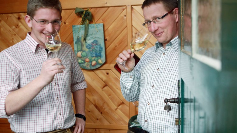 Zwei M&auml;nner in karierten Hemden trinken Wei&szlig;wein in einem holzget&auml;felten Raum.