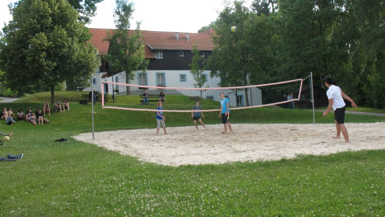 Menschen spielen Volleyball auf einem Sandplatz im Freien, umgeben von Bäumen und einem Gebäude im Hintergrund.