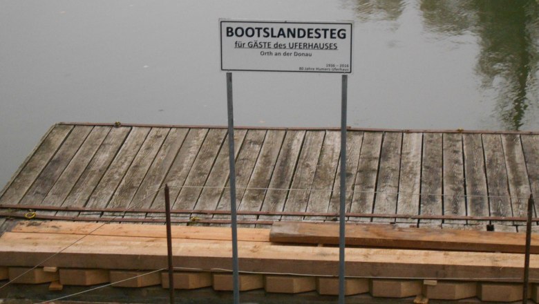Holzsteg mit Schild 'Bootslandesteg f&uuml;r G&auml;ste des Uferhauses' an einem ruhigen Gew&auml;sser.