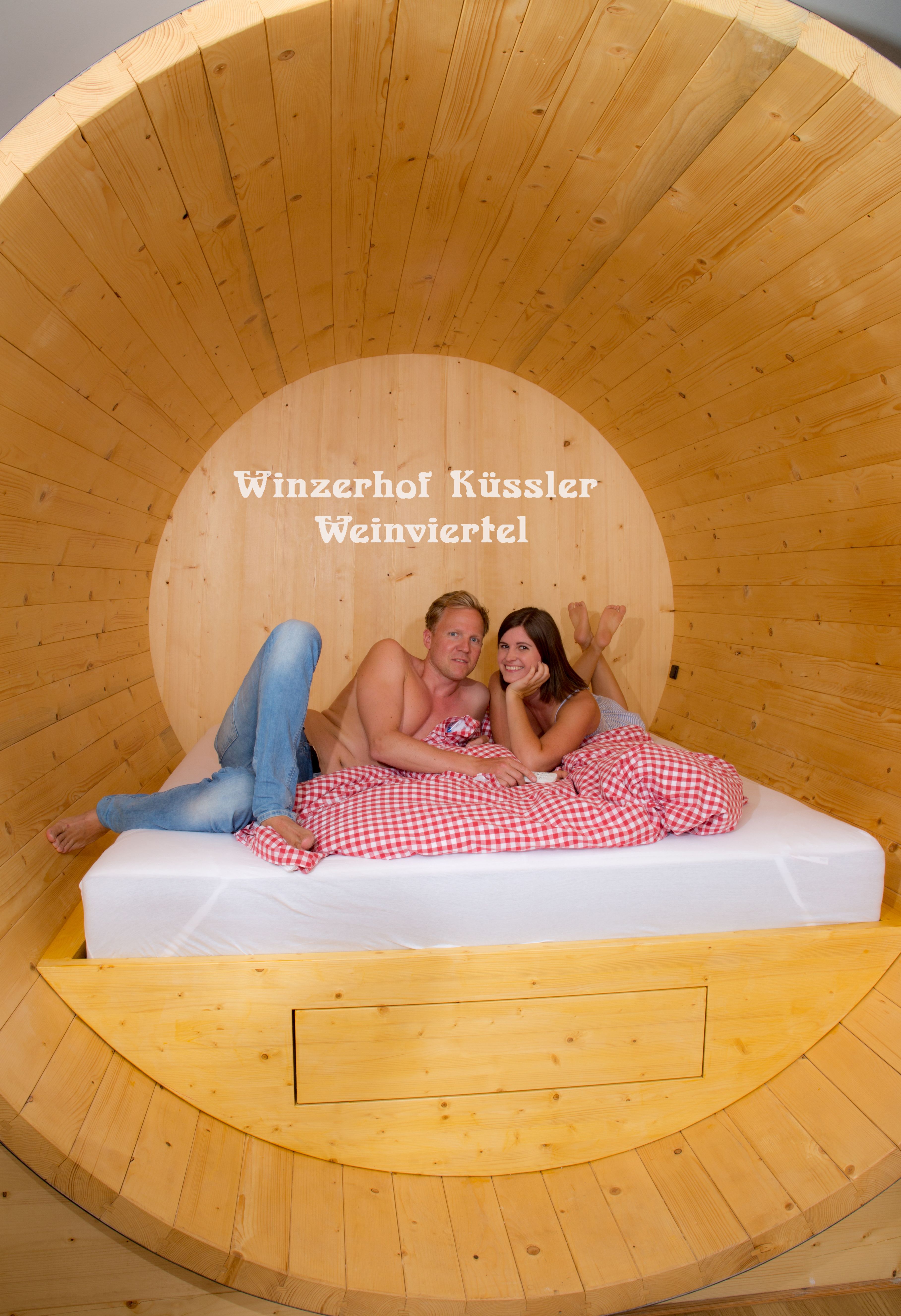 Ein Paar liegt in einem großen Holzfassbett mit rot-weiß kariertem Bettzeug. Im Hintergrund steht 'Winzerhof Küssler Weinviertel'.