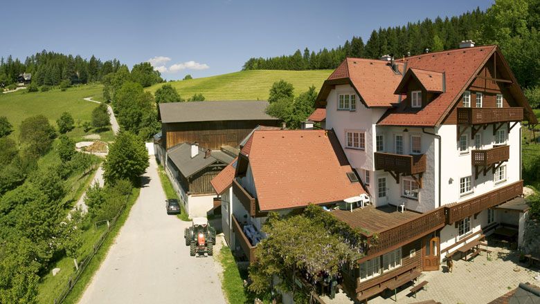 Ein Bauernhof mit roten Dächern in einer ländlichen Umgebung, umgeben von grünen Wiesen und Wäldern.