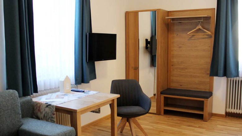 Modernes Gästezimmer mit Tisch, Stuhl, Kleiderschrank, Sofa, Fernseher und blauen Vorhängen.