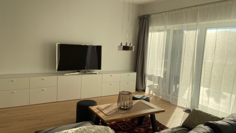 Moderner Wohnbereich mit grauem Sofa, Holztisch, Teppich, Fernseher und gro&szlig;en Fenstern mit Vorh&auml;ngen.