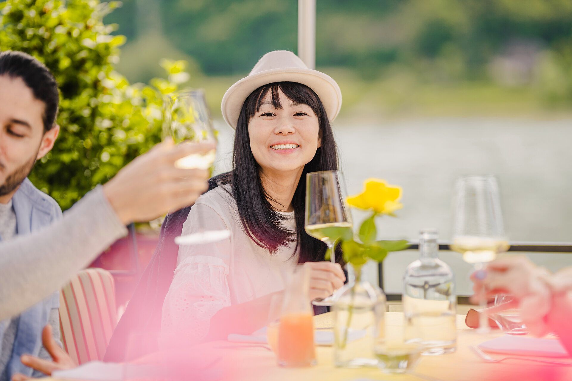 Eine Dame mit Hut sitzend an einem Tisch hält ein gefülltes Weißweinglas hoch.