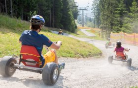 Personen fahren mit Mountaincarts einen Schotterweg hinunter, umgeben von Bäumen und einem Skilift.
