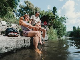 Zwei Personen sitzen auf einer Steintreppe am Flussufer und k&uuml;hlen ihre F&uuml;&szlig;e im Wasser.