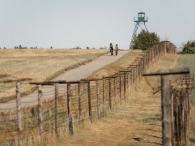 Zwei Radfahrer auf einem Weg neben einem alten Grenzzaun und einem Wachturm.