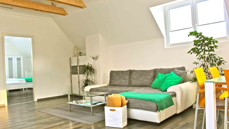 ferien-wohnen, © Frau Lendl Helles Wohnzimmer mit grauem Sofa, grünen Kissen, Glastisch und Essbereich mit gelben Stühlen.