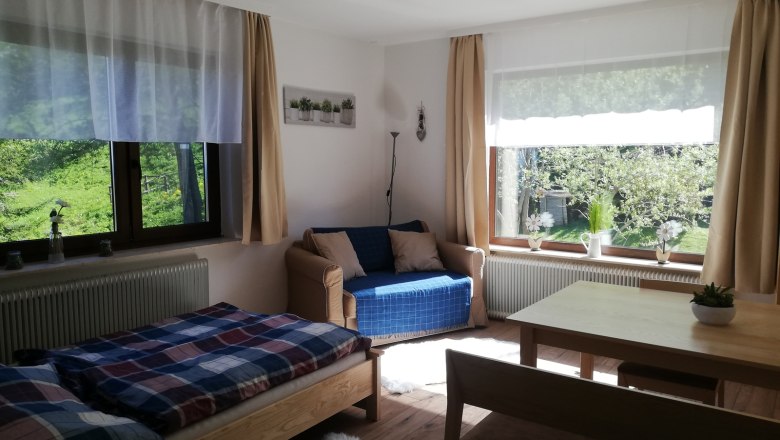 Helles Wohnzimmer mit Bett, Sofa, Tisch und großen Fenstern mit Blick ins Grüne.