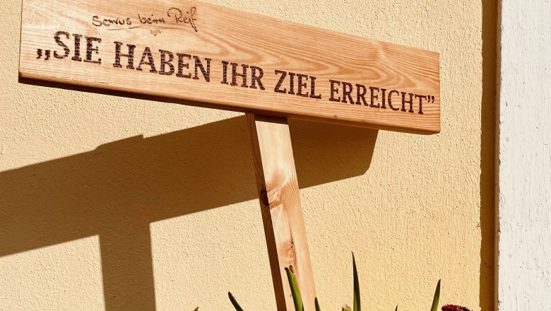 Holzschild mit der Aufschrift 'Sie haben Ihr Ziel erreicht' vor einer gelben Wand, umgeben von Blumen.