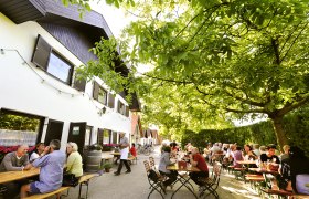 Menschen sitzen an Tischen im Freien unter B&auml;umen bei einem Heurigenbetrieb.