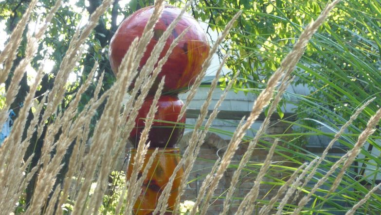 Bunte Skulptur im Garten hinter Gräsern und Pflanzen.