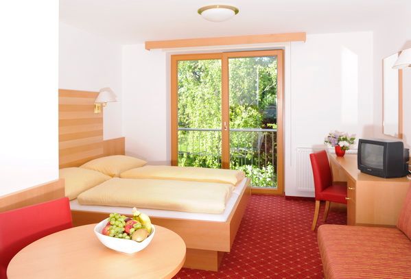 Helles Hotelzimmer mit Doppelbett, rotem Teppich, Tisch mit Obstschale und Fernseher, Blick auf grüne Bäume durch ein großes Fenster.