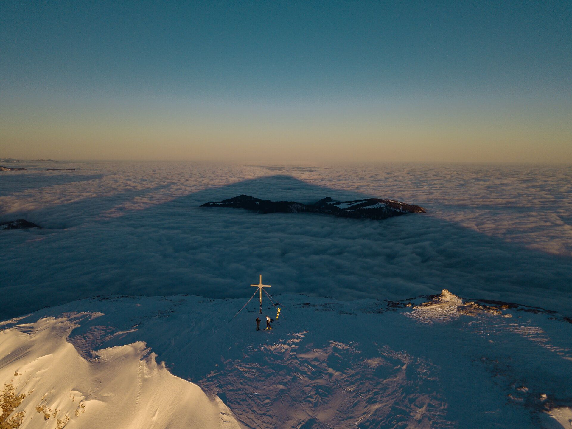 Luftaufnahme des Gipfelkreuzes am Ötscher; man sieht 2 Skitourengeher. Der Ötscher wirft einen mächtigen Schatten auf die Wolkendecke,