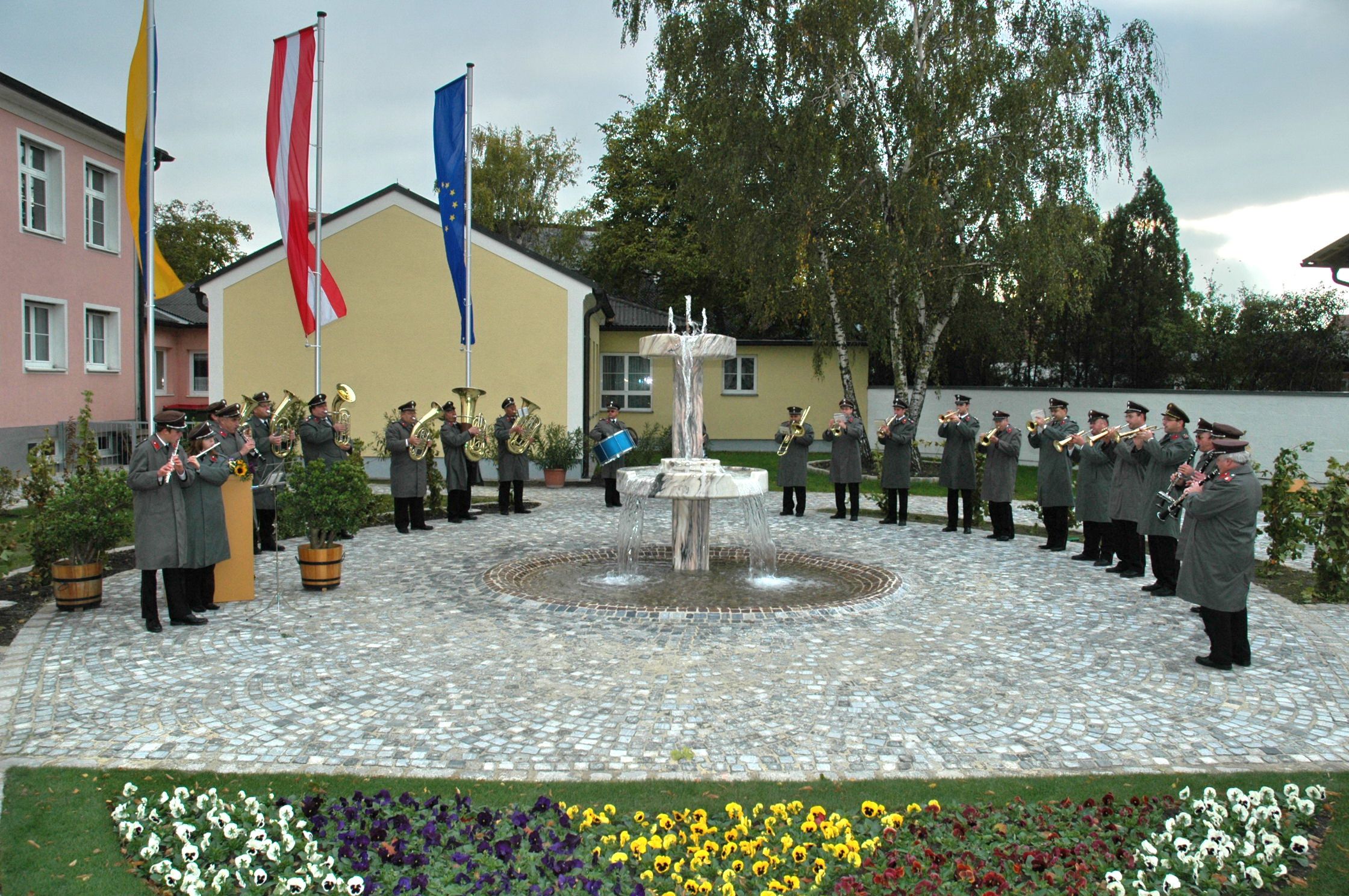 Eine Blaskapelle spielt im Freien vor einem Brunnen, umgeben von Blumen und Flaggen.