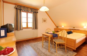 Gemütliches Gästezimmer mit Holzmöbeln, Doppelbett, kleinem Tisch und Stühlen, TV und Fenster mit Vorhängen.