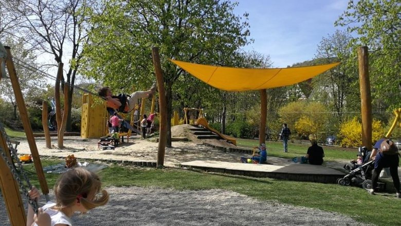 Kinder spielen auf einem Spielplatz mit Schaukeln und Kletterger&uuml;sten unter einem gelben Sonnensegel.