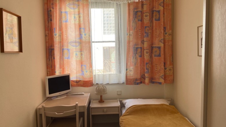 Ein kleines Einzelzimmer mit Bett, Schreibtisch, Stuhl und Fernseher. Bunte Vorh&auml;nge und eine Lampe sind ebenfalls zu sehen.