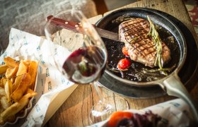 Ein Steak in einer Pfanne mit Tomaten und Rosmarin, daneben Pommes und ein Glas Rotwein auf einem Holztisch.