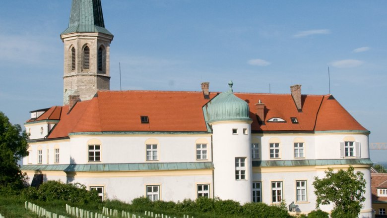 Das Schloss Gumpoldskirchen, © Schloss Gumpoldskirchen Betriebs-GmbH Schloss Gumpoldskirchen mit rotem Dach und Turm, umgeben von Weinbergen unter blauem Himmel.