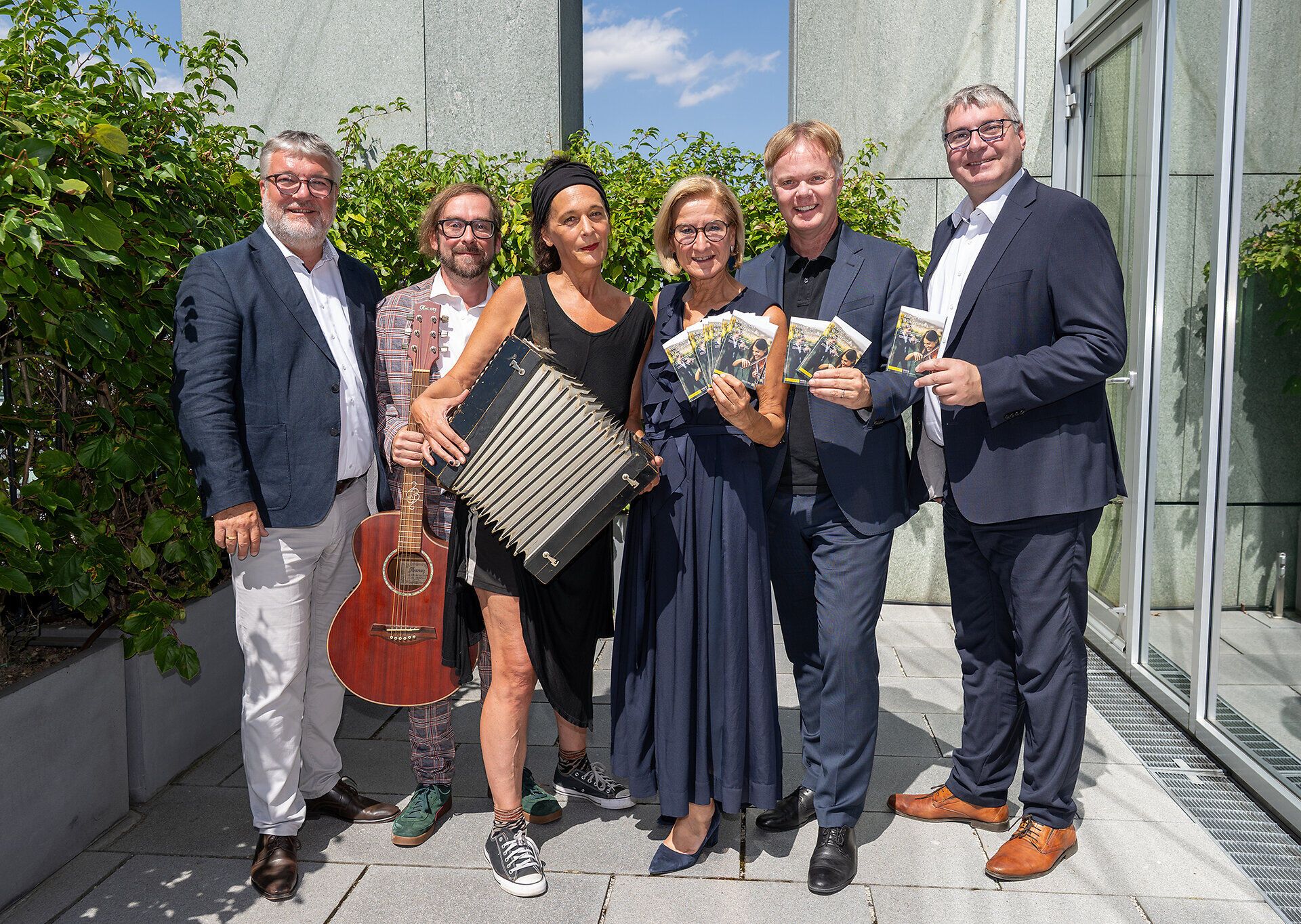 Sechs Personen stehen bei sonnigem Wetter auf einer Terrasse vor einem modernen Gebäude. Zwei Personen halten Musikinstrumente – eine Gitarre und ein Akkordeon –, drei weitere präsentieren Broschüren. Im Hintergrund sind üppige grüne Pflanzen zu sehen.