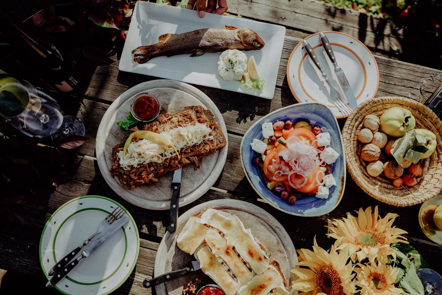 Verschiedene Gerichte auf einem Holztisch, darunter ein ganzer Fisch, Salat, belegtes Brot und Sonnenblumen.