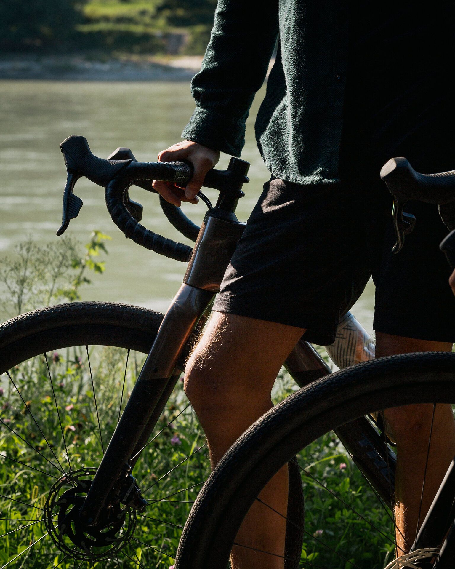 Eine Nahaufnahme von der unteren Hälfte eines Radfahrenden, der neben einem modernen Fahrrad mit Rennlenker steht. Der Hintergrund zeigt die Donau, die eine beruhigende Kulisse bietet. Das Bild fängt das Gefühl von Bewegung und sportlicher Aktivität ein, während die Person sich in einem Moment der Ruhe und Reflexion befindet.