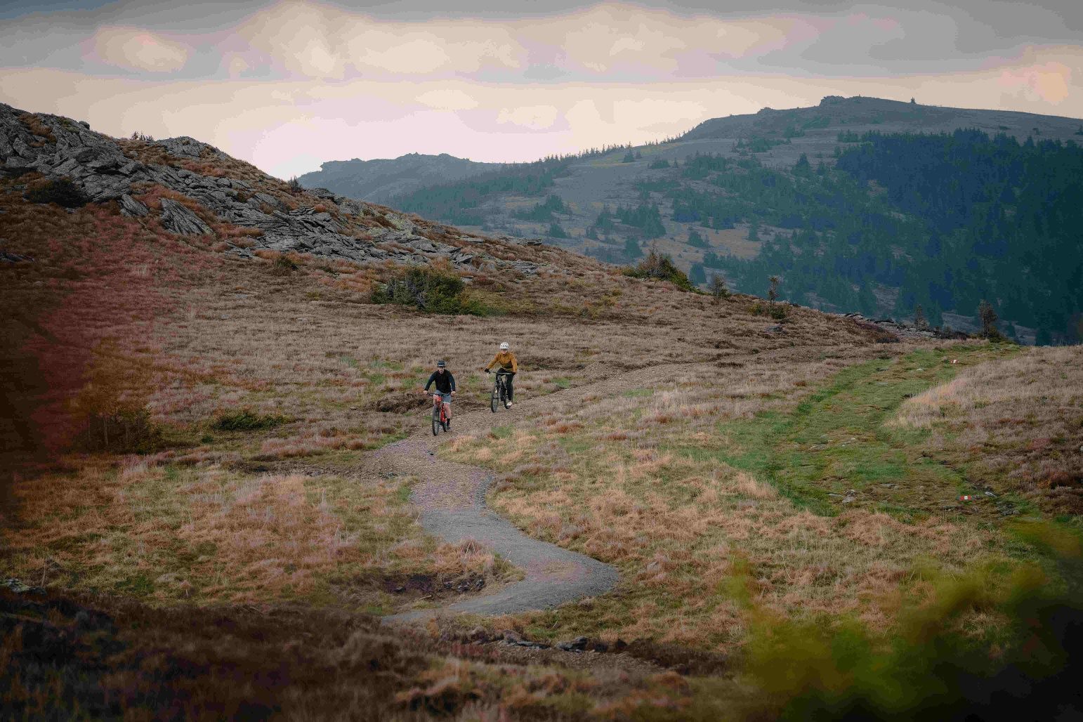 Zwei Mountainbiker fahren auf einem schmalen Pfad in einer bergigen Landschaft.