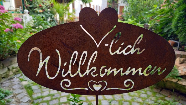 Herzlich Willkommen, © Familie Fleischmann Metallschild mit der Aufschrift 'Herzlich Willkommen' in einem Garten.