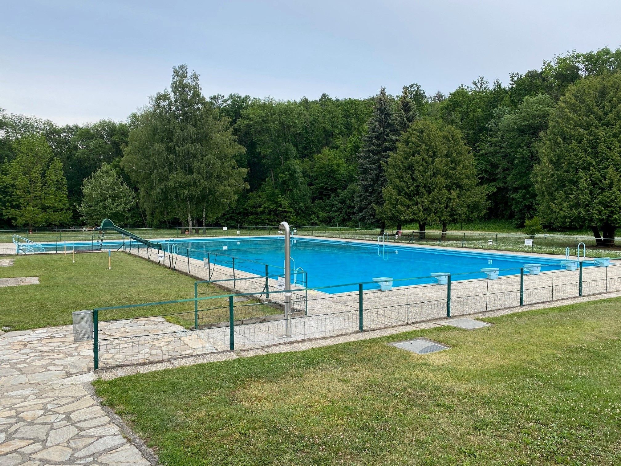 Ein leeres Freibad mit einem großen Schwimmbecken, umgeben von Bäumen und Rasenflächen.